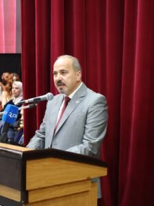 WhatsApp Image 2026-07-18 at 104005 AM 1 - عروبة الاخباري | Oroba News Man in a gray suit and red tie speaks at a wooden lectern with a microphone, red stage curtains behind him.