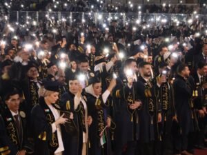 WhatsApp Image 2026-07-18 at 104005 AM 2 - عروبة الاخباري | Oroba News A large crowd of graduates in dark robes with gold trim, raising phones with flash during a ceremony.