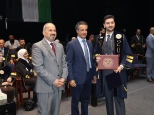 WhatsApp Image 2026-07-18 at 104005 AM 3 - عروبة الاخباري | Oroba News Graduate in a navy gown with gold trim holds a diploma while standing with two suited men at a formal ceremony, with seated attendees in the background.