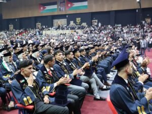 WhatsApp Image 2026-07-18 at 104005 AM - عروبة الاخباري | Oroba News Rows of graduates in dark blue gowns and mortarboards clap as a large audience watches from the stands in a large indoor venue, with Jordan flags and a portrait on the wall above.