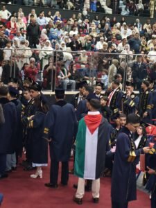WhatsApp Image 2026-07-18 at 104005 AM 4 - عروبة الاخباري | Oroba News Graduates in blue gowns stand at the front of a crowded indoor graduation ceremony, a Palestinian flag draped over one person in the center of the scene.