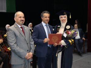 WhatsApp Image 2026-07-18 at 104005 AM 5 - عروبة الاخباري | Oroba News Graduate in cap and gown receives a diploma and bouquet from two officials on stage during a ceremony.
