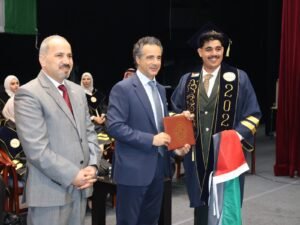 WhatsApp Image 2026-07-18 at 104005 AM 7 - عروبة الاخباري | Oroba News Graduate in cap and gown receives a diploma from officials during a graduation ceremony, with year 2020 on the sash, smiling at the camera.
