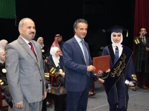 WhatsApp Image 2026-07-18 at 104006 AM 1 - عروبة الاخباري | Oroba News Graduate in a navy cap and gown with a white hijab receives a diploma from a suited man on stage.