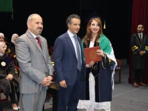 WhatsApp Image 2026-07-18 at 104006 AM 2 - عروبة الاخباري | Oroba News Graduate in cap and gown holding a diploma, flanked by two men in suits on stage during a ceremony