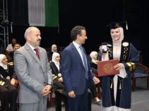 WhatsApp Image 2026-07-18 at 104006 AM 3 - عروبة الاخباري | Oroba News Graduate woman in blue and gold gown and hijab holds up a photo on her phone while receiving a diploma on stage from two men in suits.