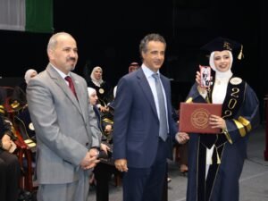 WhatsApp Image 2026-07-18 at 104006 AM - عروبة الاخباري | Oroba News Graduation scene: a smiling graduate in a hijab and cap holds a diploma and a red phone, shaking hands with two men in suits at a ceremony.