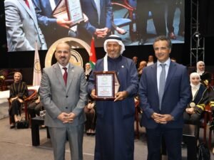 WhatsApp Image 2026-07-18 at 104006 AM 4 - عروبة الاخباري | Oroba News Three men in formal attire on a stage during an award ceremony; the center man in traditional dress holds a framed plaque while others stand beside him, with a Jordanian flag in the background.