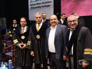 WhatsApp Image 2026-07-18 at 104006 AM 5 - عروبة الاخباري | Oroba News Group of men in dark graduation gowns with gold trim posing on stage at a ceremony, a banner reading 'Queen Noor Aviation University College' hangs behind them.