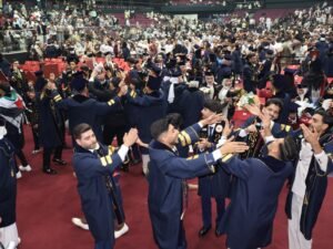 WhatsApp Image 2026-07-18 at 104006 AM 6 - عروبة الاخباري | Oroba News Graduates in navy gowns with gold trim cheer and toss hats on a red-carpeted floor as a crowded arena watches.