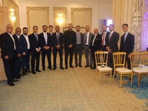 WhatsApp Image 2026-07-18 at 112231 AM 1 - عروبة الاخباري | Oroba News Fifteen men in suits posing together for a group photo in an ornate, gold-accented banquet hall.