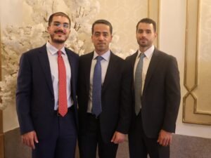 WhatsApp Image 2026-07-18 at 112231 AM 2 - عروبة الاخباري | Oroba News Three men in suits posing together at a formal indoor event, with decorative wallpaper and a white floral display behind them.
