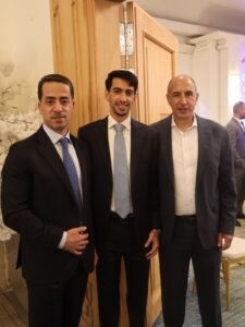 WhatsApp Image 2026-07-18 at 112231 AM 3 - عروبة الاخباري | Oroba News Three men in suits standing together at an indoor event near a wooden door.