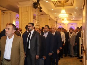 WhatsApp Image 2026-07-18 at 112232 AM 2 - عروبة الاخباري | Oroba News Formal group of men in suits walking single-file through a ballroom lined with columns and chandeliers at an event venue.