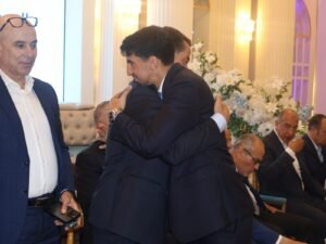 WhatsApp Image 2026-07-18 at 112232 AM 4 - عروبة الاخباري | Oroba News Two men in dark suits hug at a formal event, with others seated and white floral arrangements in the background.