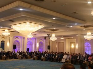 WhatsApp Image 2026-07-18 at 112233 AM 1 - عروبة الاخباري | Oroba News Elegant event hall with crystal chandeliers and rows of men in suits seated along the perimeter of a luxurious room.