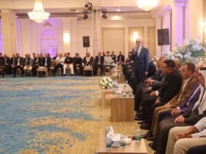 WhatsApp Image 2026-07-18 at 112233 AM 3 - عروبة الاخباري | Oroba News Man in a blue suit speaks into a microphone to a formal audience in an ornate ballroom with chandeliers and white floral decor.
