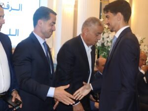 WhatsApp Image 2026-07-18 at 112233 AM - عروبة الاخباري | Oroba News Two businessmen in dark suits shake hands at a formal event as others look on in a bright room with floral decor.