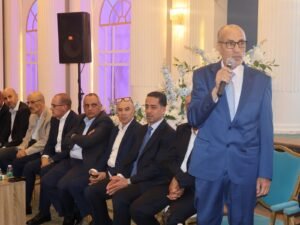 WhatsApp Image 2026-07-18 at 112233 AM 4 - عروبة الاخباري | Oroba News Man in a blue suit speaks into a microphone at a formal event, with seated attendees in suits and a white floral backdrop.