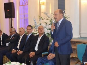 WhatsApp Image 2026-07-18 at 112233 AM 5 - عروبة الاخباري | Oroba News Man in a blue suit stands and speaks into a microphone at a formal indoor event; men in suits sit beside him with a floral backdrop behind them.