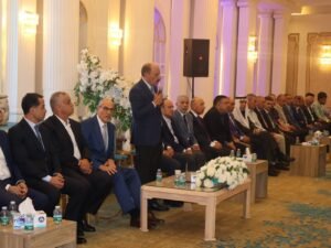 WhatsApp Image 2026-07-18 at 112233 AM 6 - عروبة الاخباري | Oroba News Man in a blue suit speaks into a handheld microphone while standing among seated men in formal attire at a ceremonial event in a grand columned hall with white flowers behind him.