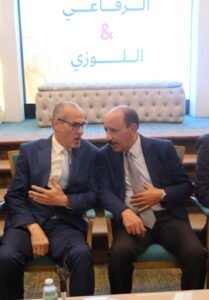 WhatsApp Image 2026-07-18 at 112234 AM 1 - عروبة الاخباري | Oroba News Two men in dark suits sit close together, mid-conversation, in a formal setting with an illuminated Arabic sign behind them.