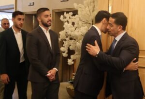 WhatsApp Image 2026-07-18 at 112234 AM 3 - عروبة الاخباري | Oroba News Two men in dark suits share a hug in a formal lobby while others wait nearby.