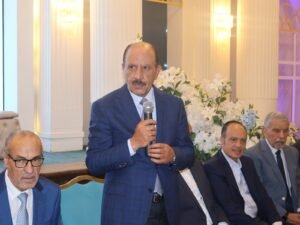 WhatsApp Image 2026-07-18 at 112234 AM - عروبة الاخباري | Oroba News Middle-aged man in a blue suit speaks into a microphone at a formal event, with other suited men seated beside him and white floral arrangements in the background.
