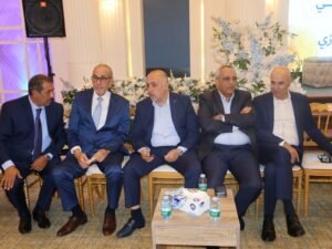 WhatsApp Image 2026-07-18 at 112235 AM - عروبة الاخباري | Oroba News Five men in dark suits sit in a row on gold chairs at a formal event, chatting with a floral backdrop behind them.