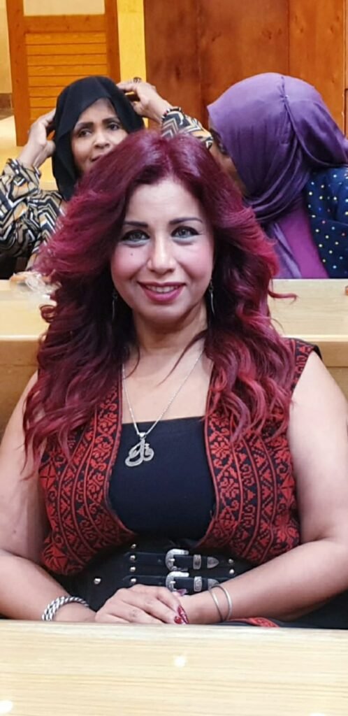 WhatsApp Image 2026-07-19 at 40022 PM3 - عروبة الاخباري | Oroba News Front-facing woman with reddish‑purple hair, wearing a black top and red patterned vest, sits at a wooden bench with a silver necklace and belt; two other women in hijabs sit behind her in a room.