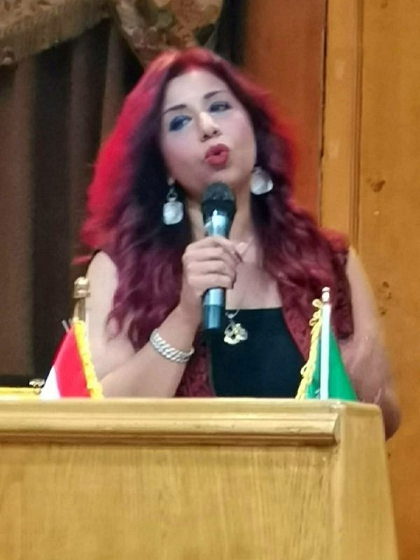 Woman with red hair singing into a microphone at a wooden podium, with two small flags (red-and-white and green) displayed on the front.