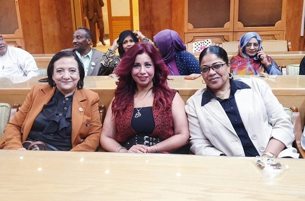 Three women sit in a wooden conference room, posing for the camera in the front row with others behind them.