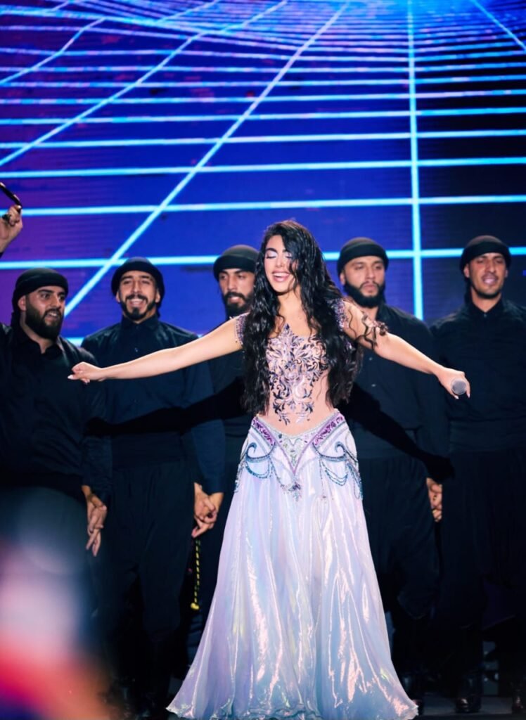 WhatsApp Image 2026-07-19 at 60539 PM - عروبة الاخباري | Oroba News Female singer in a lavender embellished gown with arms outstretched on stage, backed by dancers in black with a blue grid backdrop.