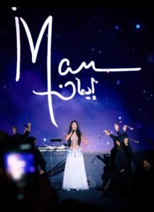 WhatsApp Image 2026-07-19 at 60540 PM - عروبة الاخباري | Oroba News Female singer in a silver gown performing on stage, backed by a large white script logo on a blue-lit backdrop with dancersaround her.