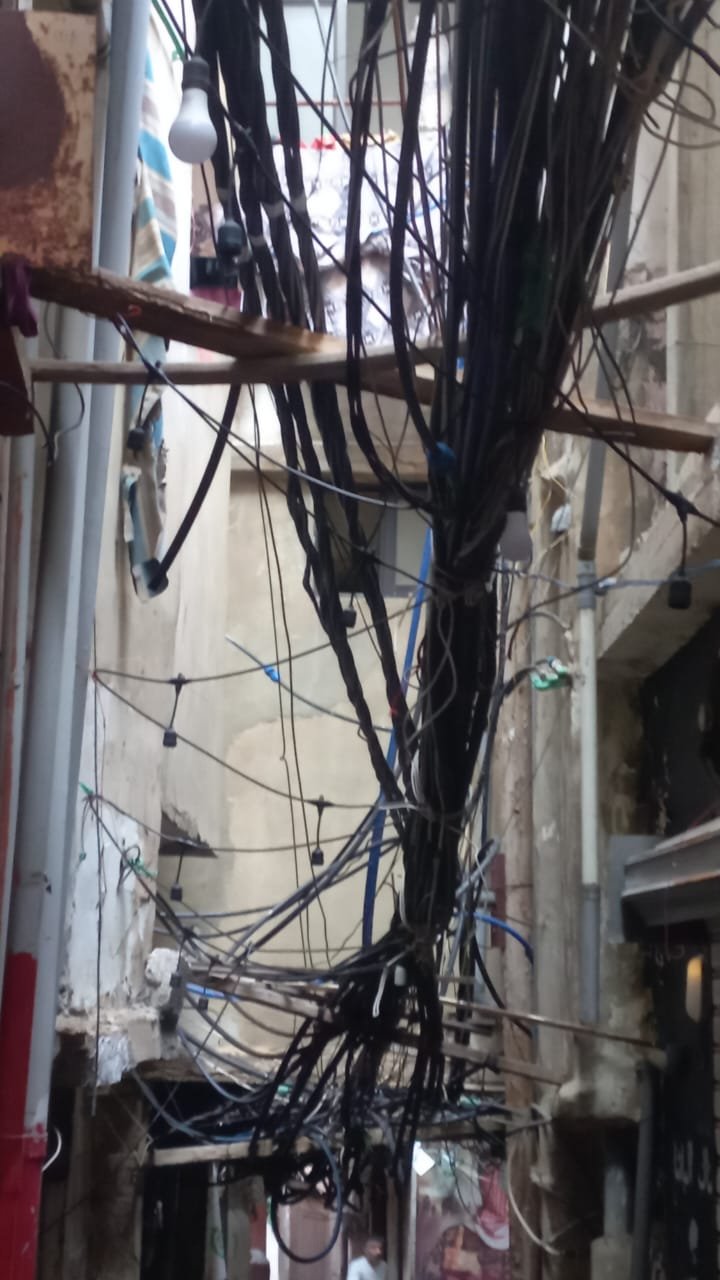 WhatsApp Image 2026-07-21 at 61530 PM Narrow alley cluttered with a tangled mass of electrical cables and a single hanging light bulb.