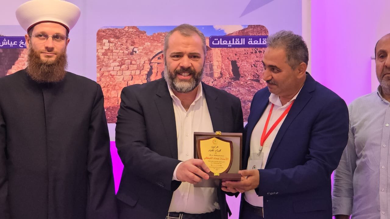 WhatsApp Image 2026-07-22 at 120120 PM 2 Three men at an award ceremony: a bearded man in a black suit receives a wooden plaque from a colleague in a navy blazer, while another man in a dark suit watches on the left; purple lighting and a banner backdrop are visible behind them.