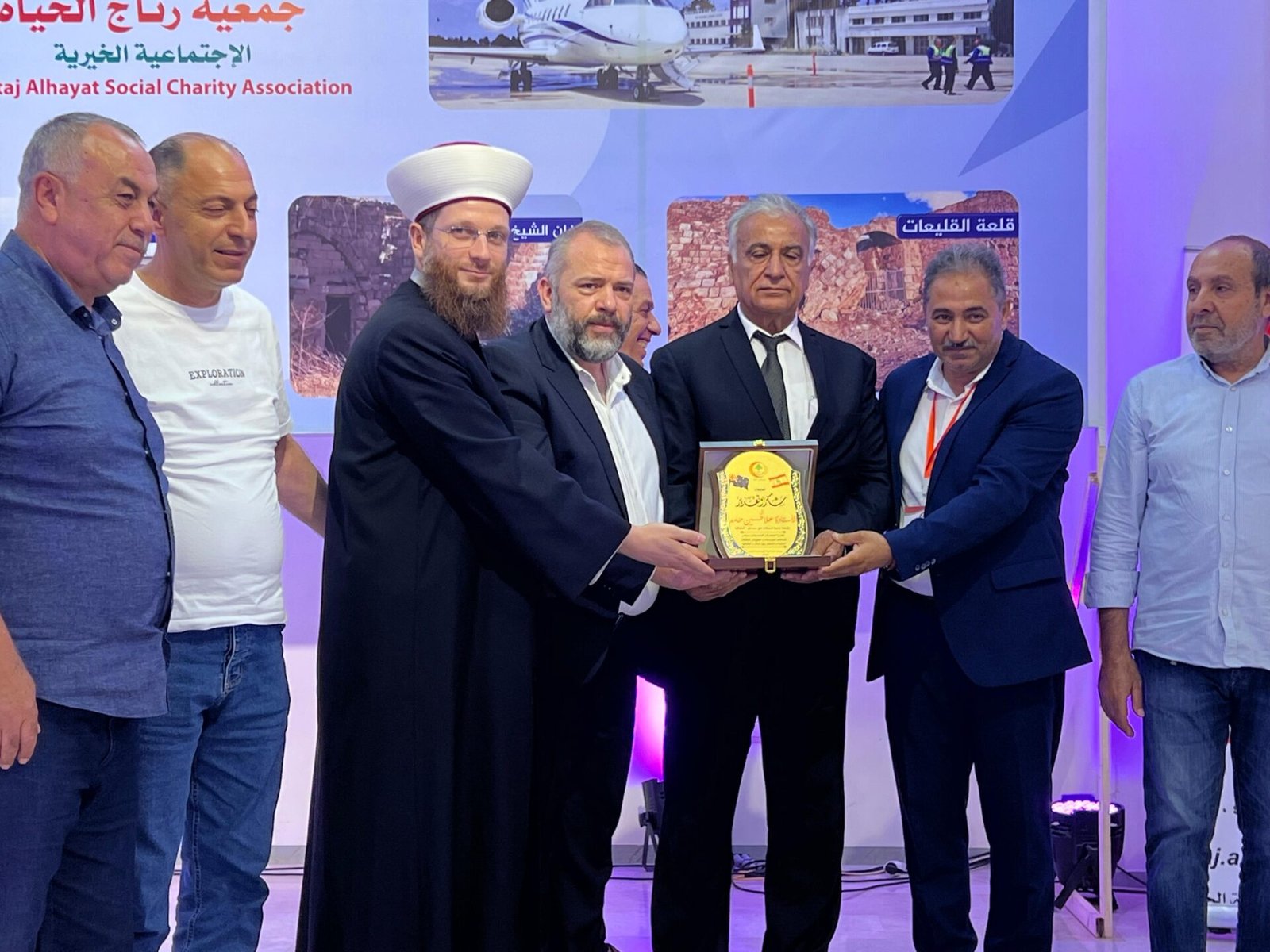 WhatsApp Image 2026-07-22 at 120121 PM 4 Group of men at an award ceremony; a man in a white turban and black robe presents a plaque to a man in a suit while others look on.