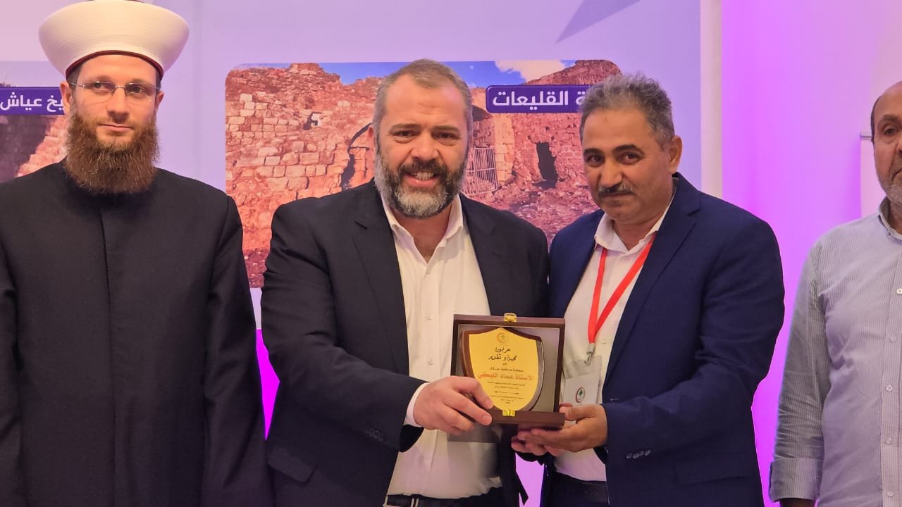 WhatsApp Image 2026-07-22 at 120121 PM Group of four men in suits on stage; center-left man hands a wooden plaque to the man on his right during an award ceremony, with a bearded cleric on the far left.