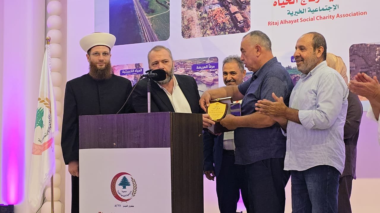 WhatsApp Image 2026-07-22 at 120123 PM 1 Group of men on a stage during a charity event; one man speaks at a podium as another hands him a shield-shaped award, while others applaud in the background.