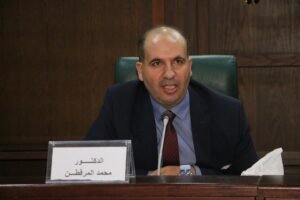 WhatsApp Image 2026-07-22 at 125638 AM - عروبة الاخباري | Oroba News Man in a suit speaks into a microphone at a panel, seated behind a wooden desk with an Arabic nameplate in front.