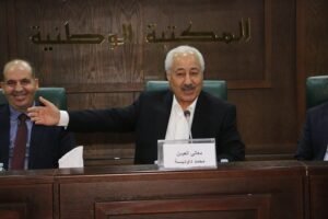 WhatsApp Image 2026-07-22 at 125641 AM - عروبة الاخباري | Oroba News Middle-aged man in a suit speaks at a panel, gesturing with his left hand, in a wood-panelled room with Arabic script behind him.