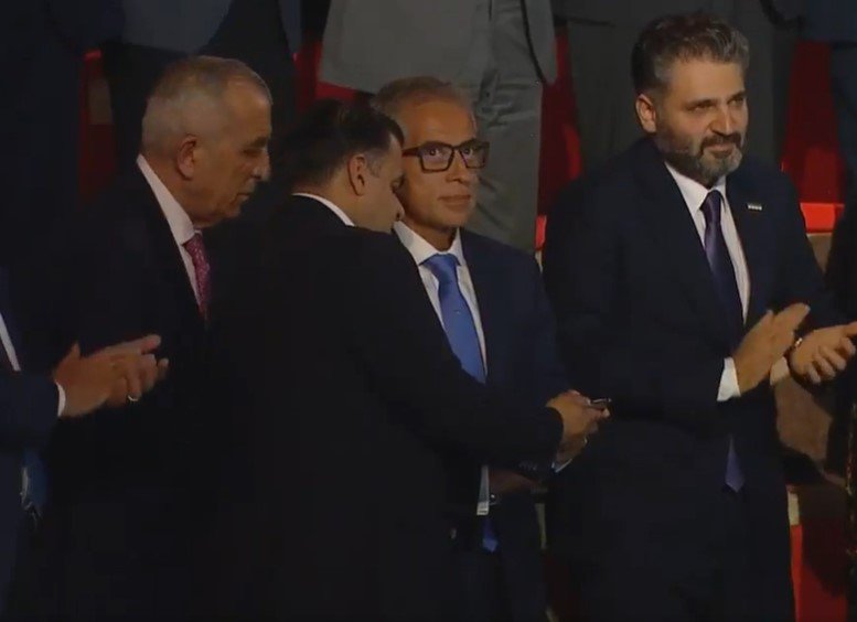 Group of suited men at a formal event; two men shake hands while others applaud nearby.