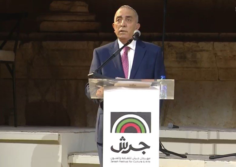 Man in a navy suit and pink tie speaks at a podium with Jerash Festival logo.