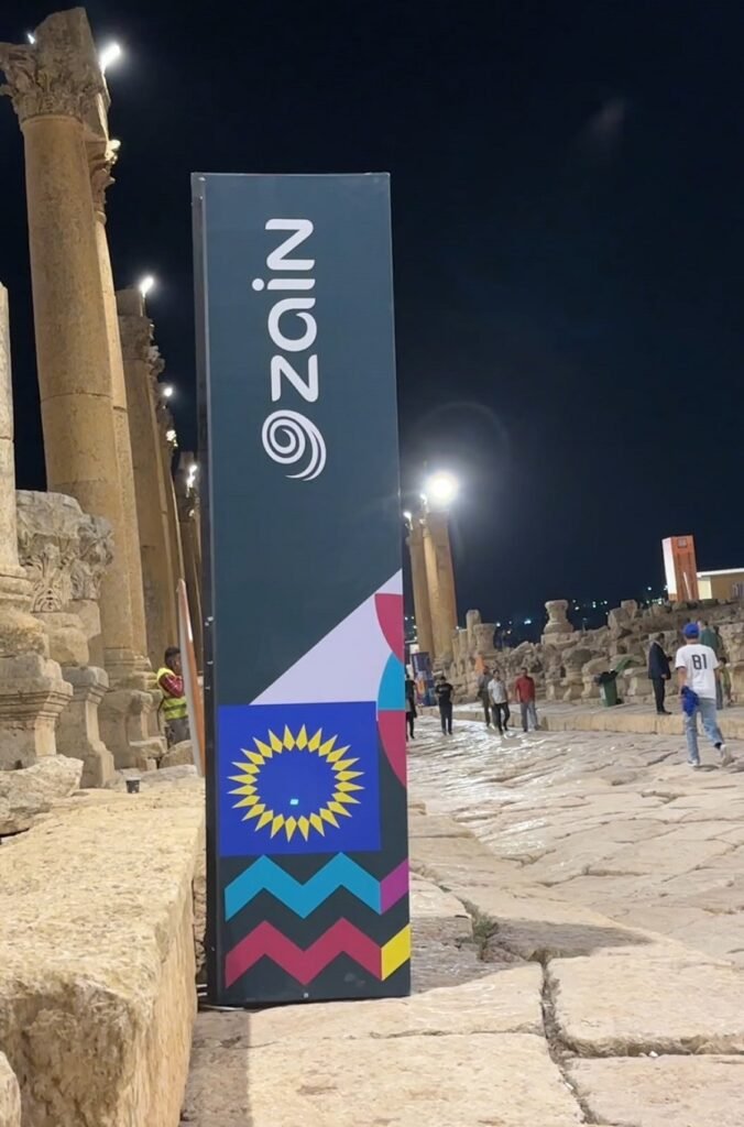 Tall vertical banner with 'Zain' logo beside ancient stone columns at night; colorful geometric design at the base.
