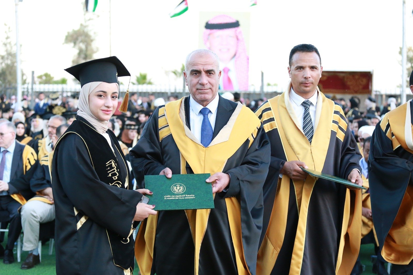 WhatsApp Image 2026-07-23 at 62959 PM Graduate woman in a hijab and cap receives a diploma from a faculty member at a graduation ceremony, with other graduates in regalia in the background.