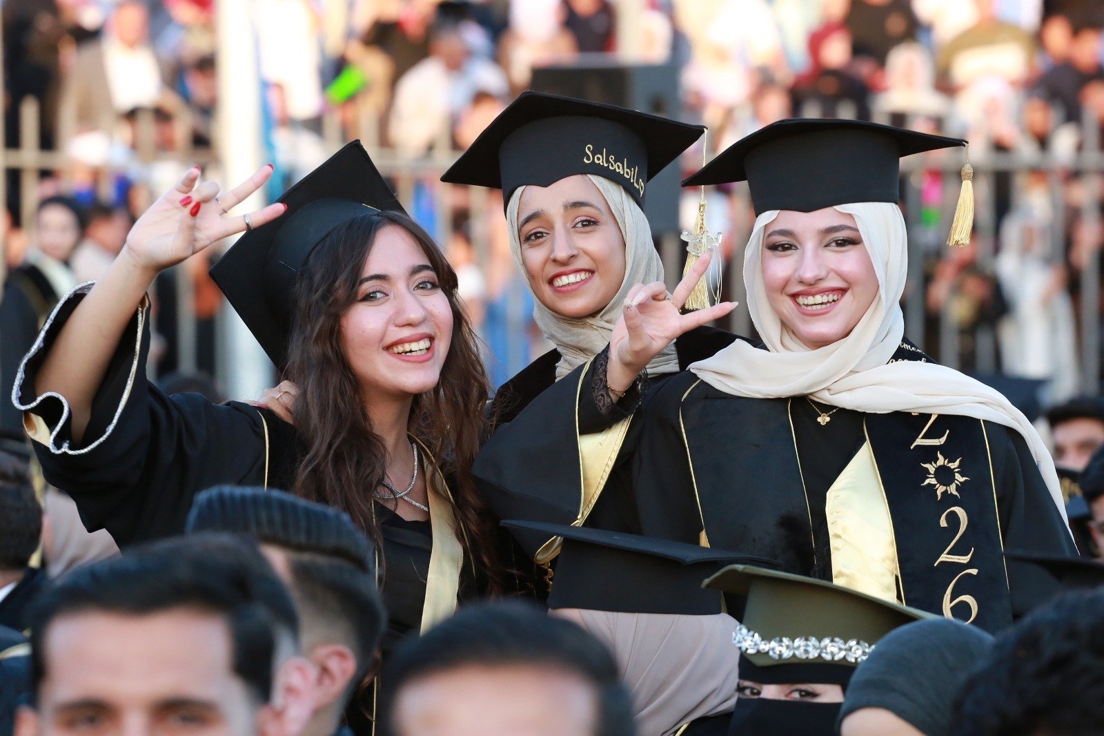 WhatsApp Image 2026-07-23 at 62959 PM1 Three graduates in black gowns and caps pose for a smiling photo at graduation, one making a peace sign with fingers visible on the left woman’s hand as a crowd looks on behind them.