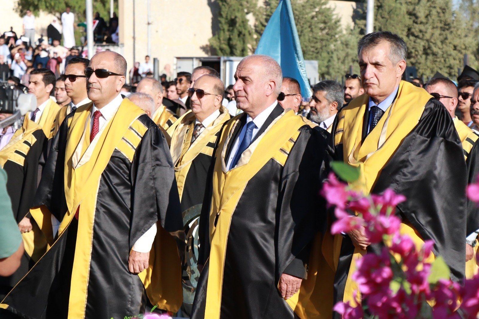 WhatsApp Image 2026-07-23 at 63000 PM Men in black and gold academic robes march at an outdoor ceremony with spectators in the background and pink flowers in the foreground.
