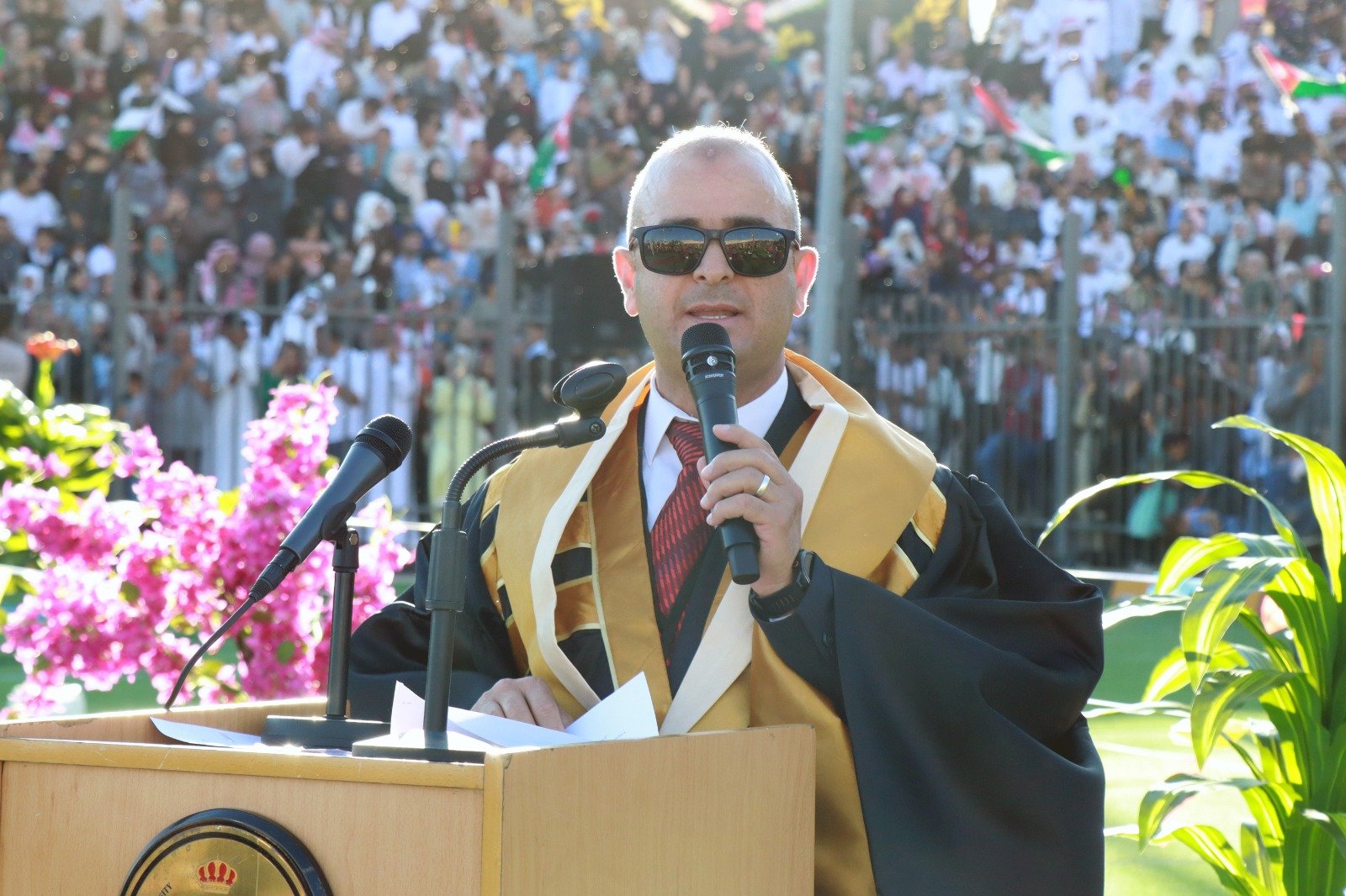 WhatsApp Image 2026-07-23 at 63000 PM2 Speaker in a black and gold graduation gown at a podium, sunglasses on, holding a microphone with a crowd behind him during an outdoor ceremony.
