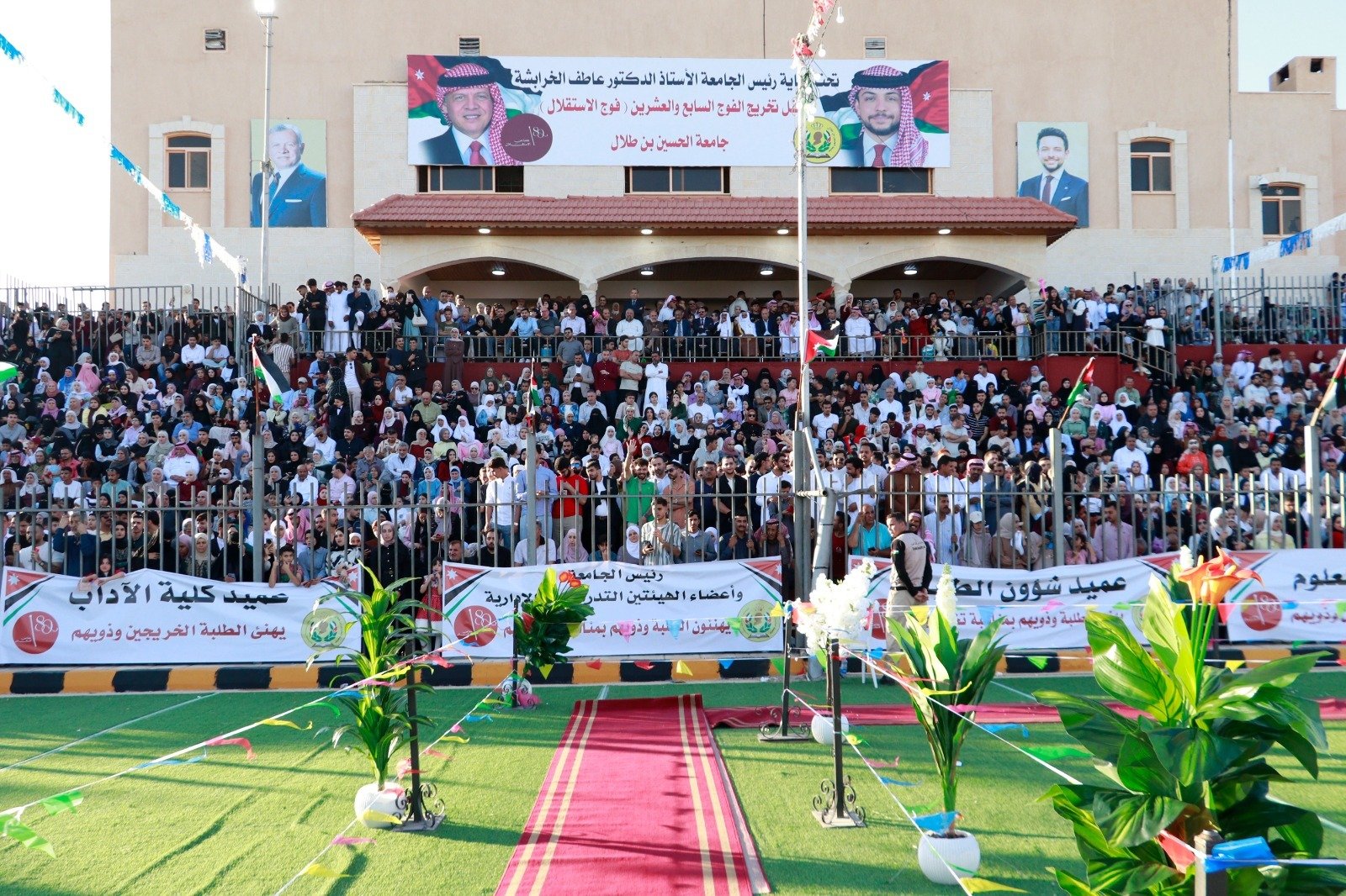 WhatsApp Image 2026-07-23 at 63001 PM Large crowd gathered in stadium stands for a ceremony with banners and flags overhead, front row banners in Arabic.