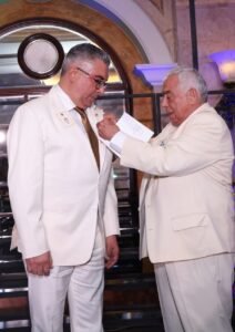 WhatsApp Image 2026-07-26 at 15355 PM - عروبة الاخباري | Oroba News Two men in white suits at a formal event; one pins a badge onto the other’s lapel during a ceremony on a staircase with an arched window behind them.